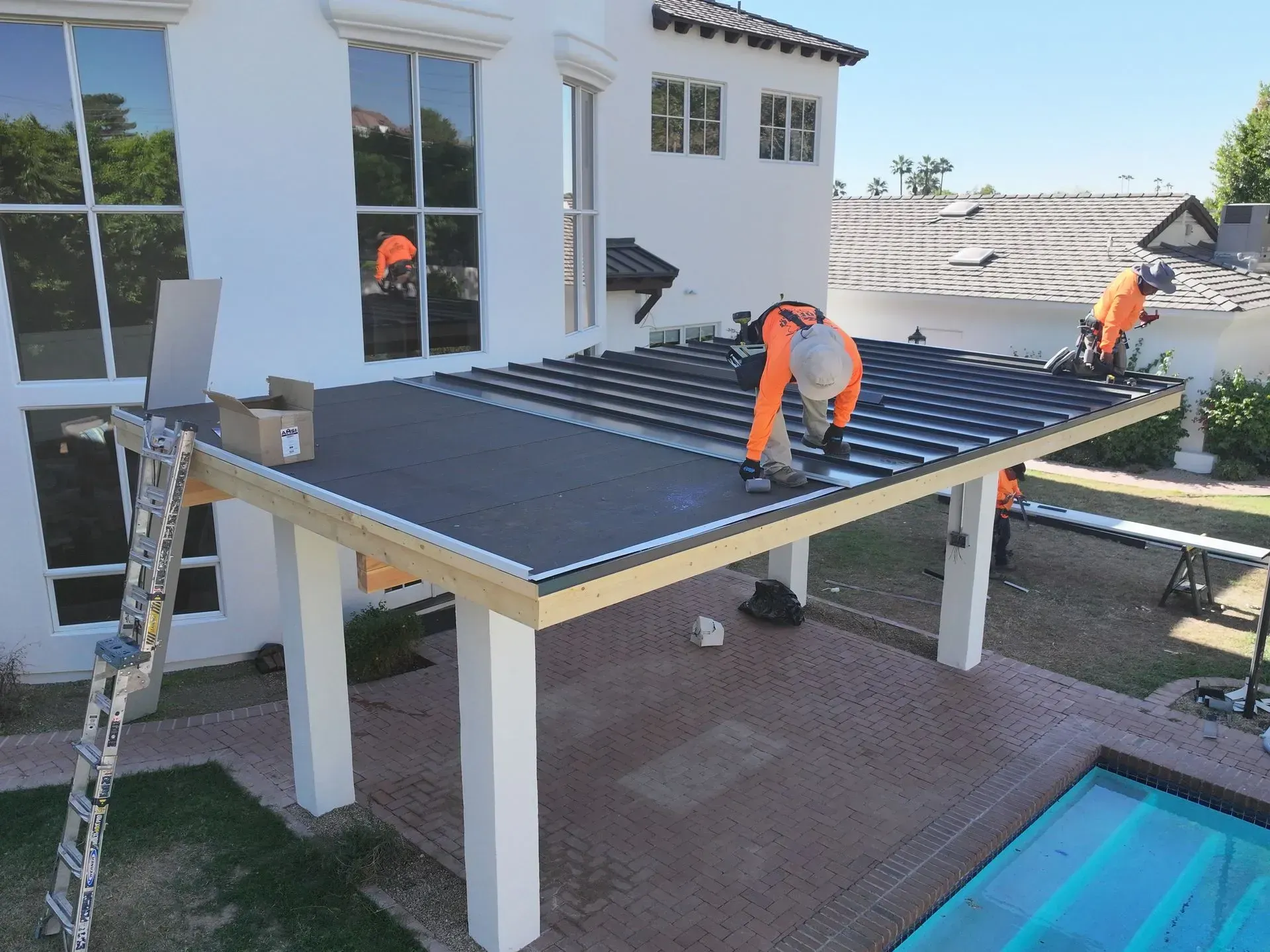 Workers installing a black pergola roof over a patio beside a house and pool