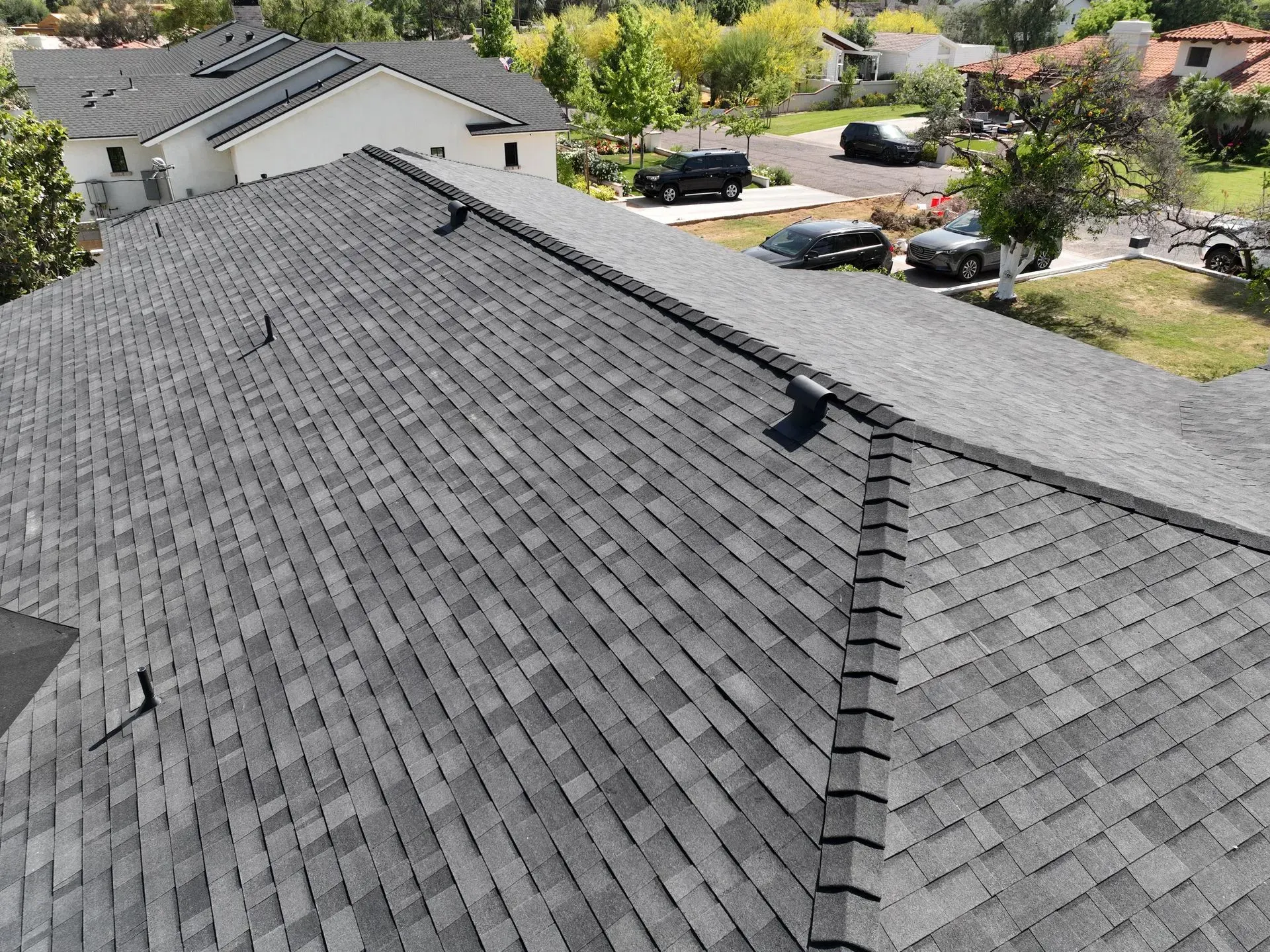 Rooftop view of gray shingles with a ridge vent, overlooking trees and parked cars in a suburban neighborhood