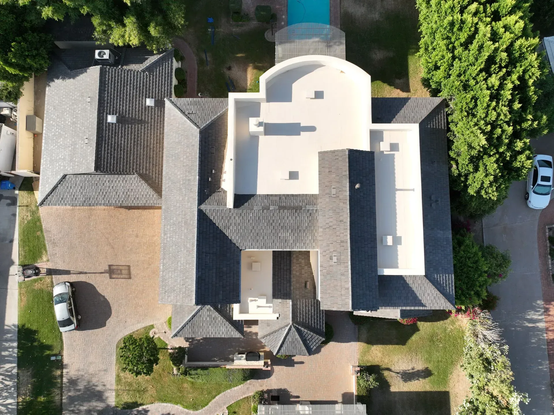 Aerial view of a house roof with driveway, trees, and parked cars around it.