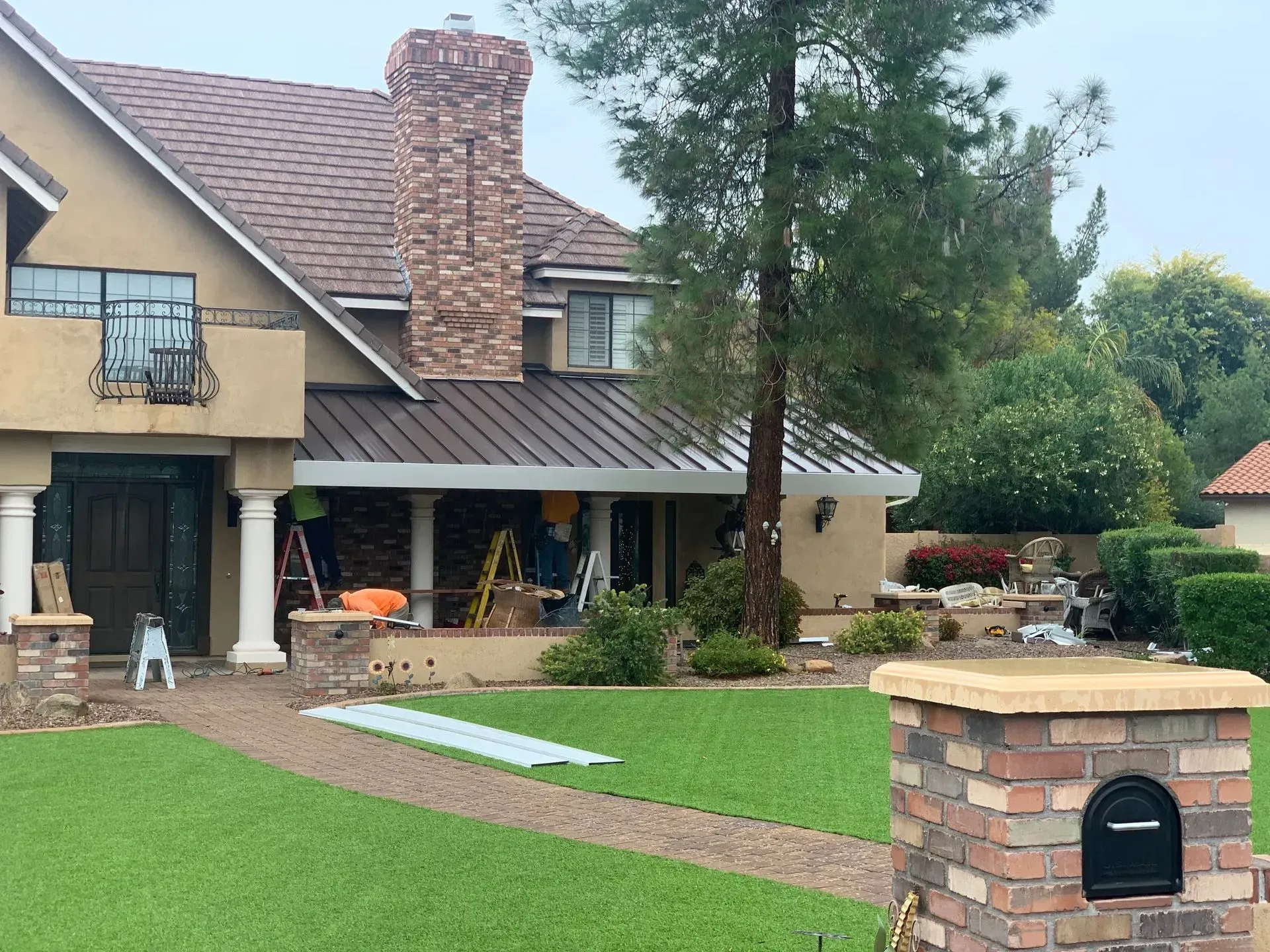 Suburban house with green lawn, stone chimney, covered patio, and brick mailbox under a cloudy sky
