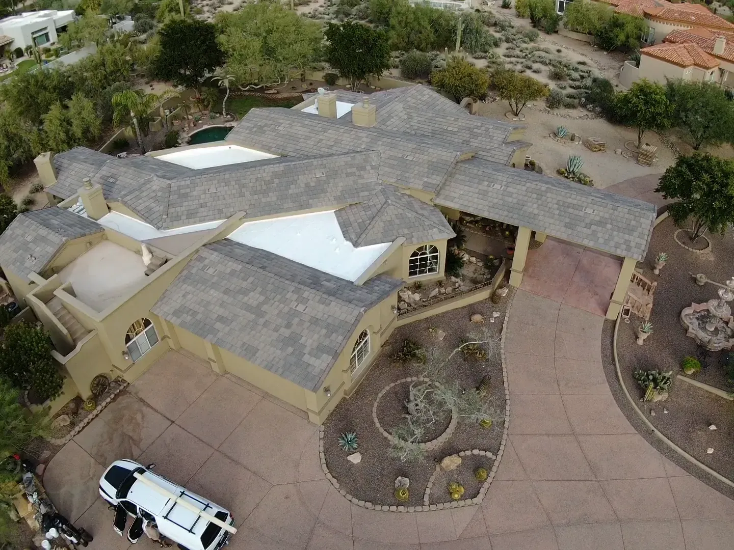 Aerial view of a beige house with a gray roof, curved driveway, and landscaped yard with a white vehicle