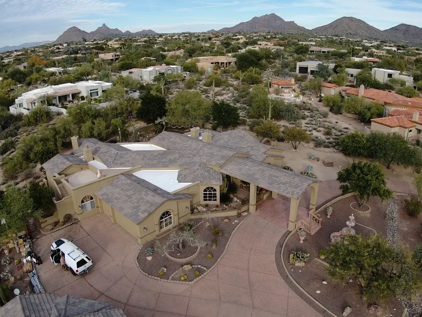 Aerial view of a tan house with a driveway, trees, and desert mountains in the background
