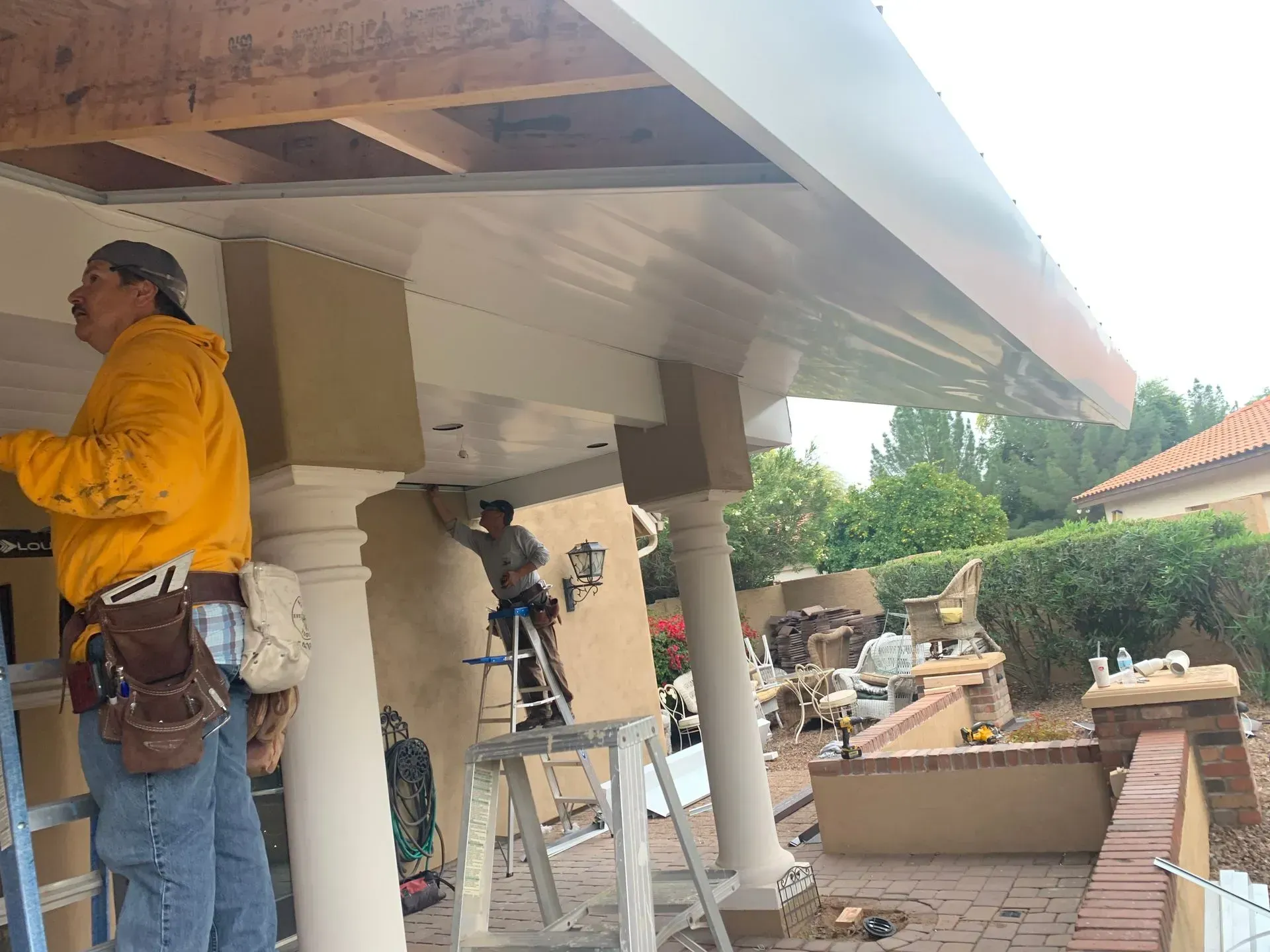 Workers painting a covered porch with white columns, ladders, and tools on site