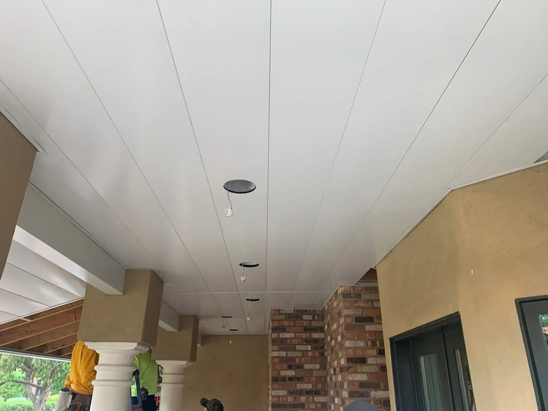 Covered outdoor walkway with white ceiling panels, tan walls, brick column, and hanging lights