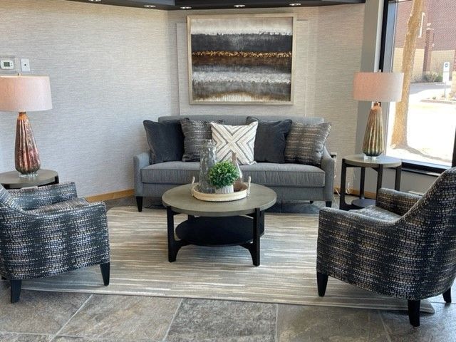 Lounge area with gray sofa, patterned armchairs, round coffee table, lamps, artwork, and a rug.