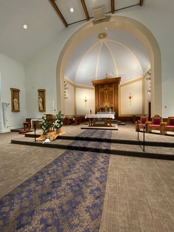 Interior of a church: Altar with wooden backdrop, blue carpet runner, beige walls, and arched ceiling.
