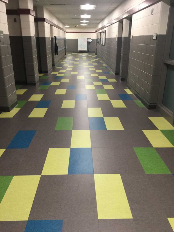Hallway with checkered floor of gray, blue, green, and yellow squares. Gray walls, doors, and overhead lights.