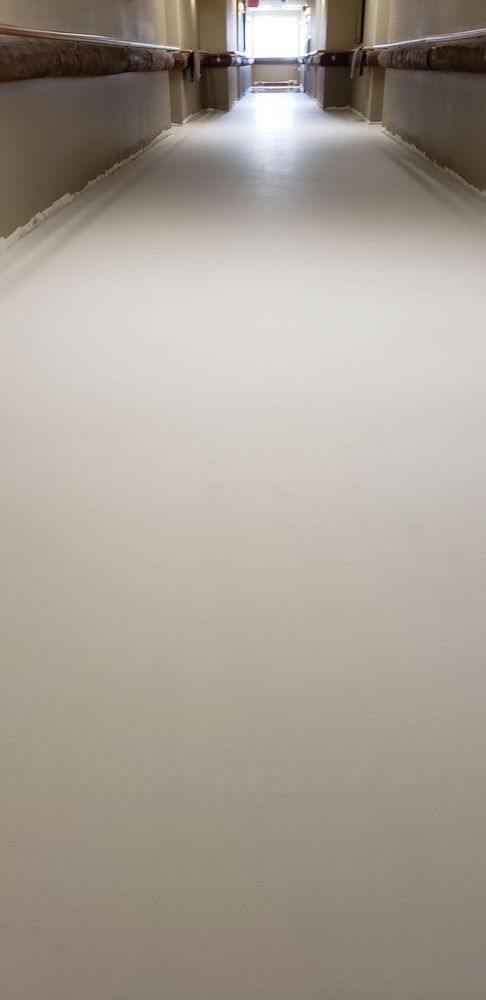 A long, empty hallway with a bright light at the end. Walls are beige, floor is white.