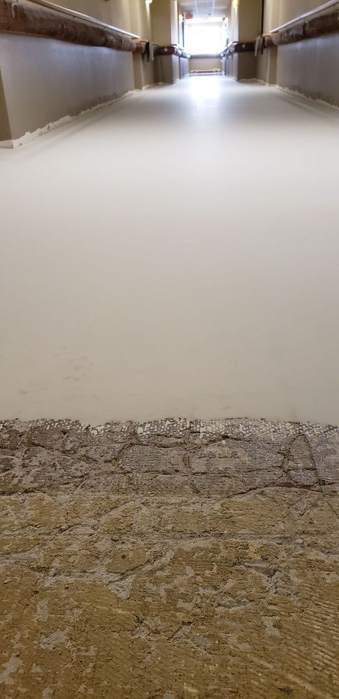 An empty, long, white corridor with a weathered stone floor. Beams and windows are at the end.