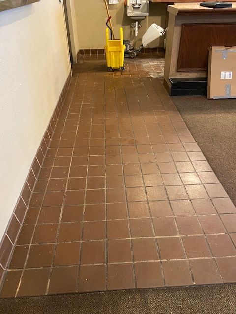 Brown tiled floor being cleaned with a yellow bucket and mop; cream wall on the left, brown counter on the right.