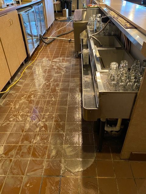 Wet restaurant floor with water and cleaning solution, near a bar with glasses and a sink.