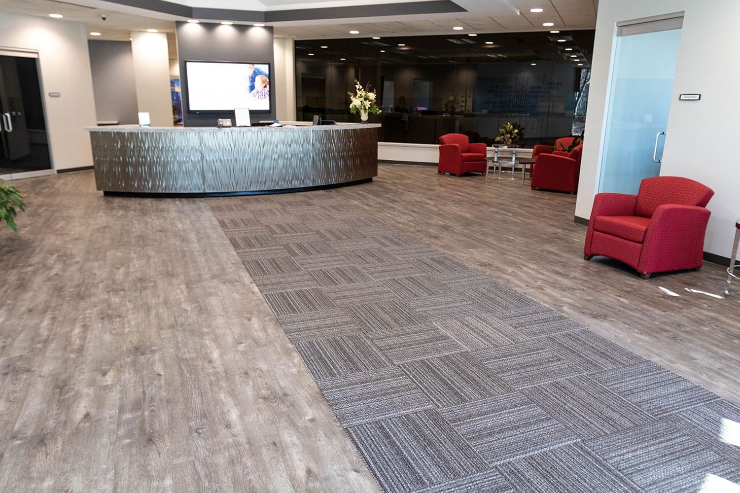 Lobby with gray and wood-look flooring, curved reception desk, red armchairs, and a TV screen.