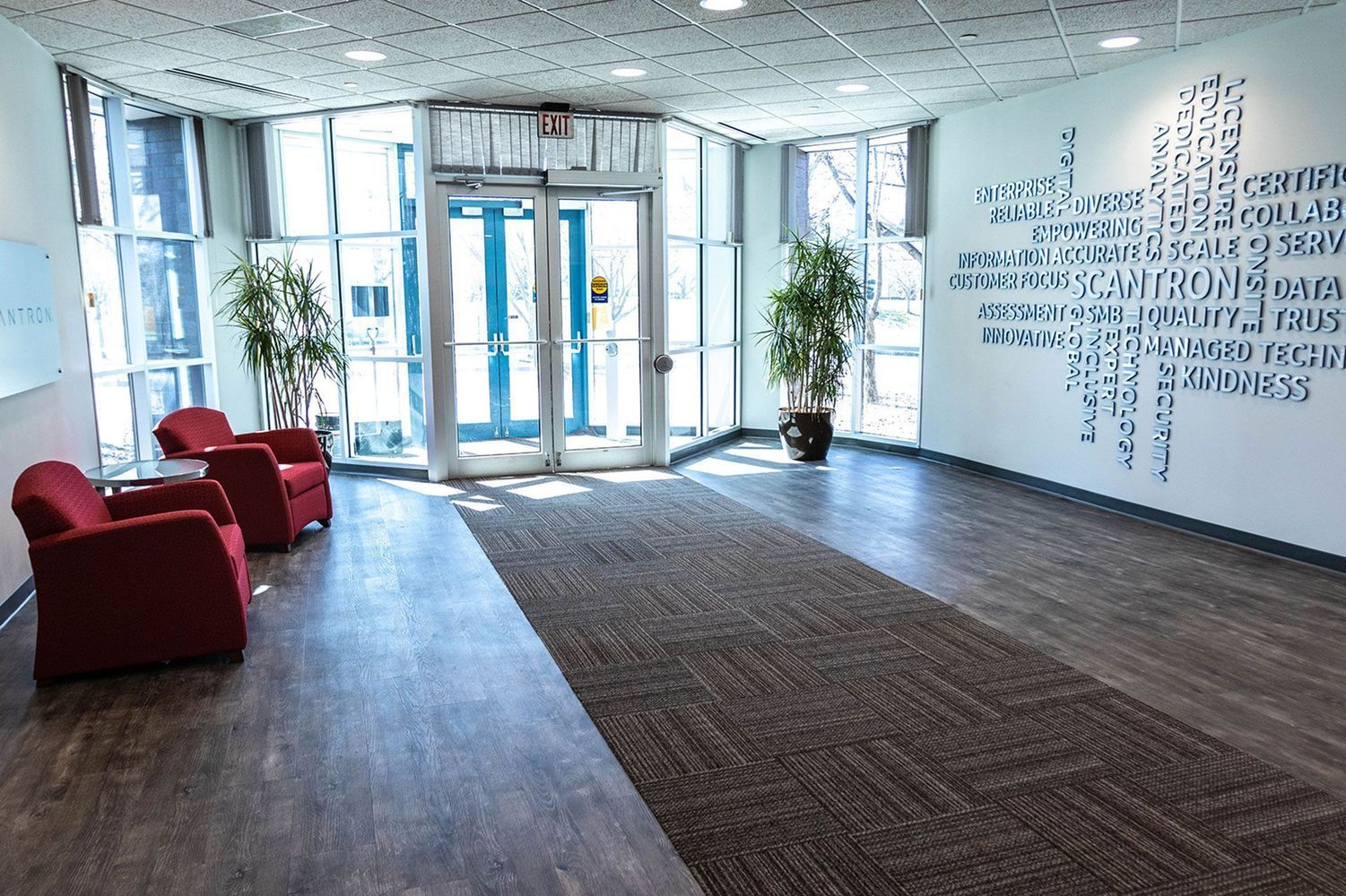 Lobby with red armchairs, indoor plants, and a sign with text. Double glass doors lead outside.