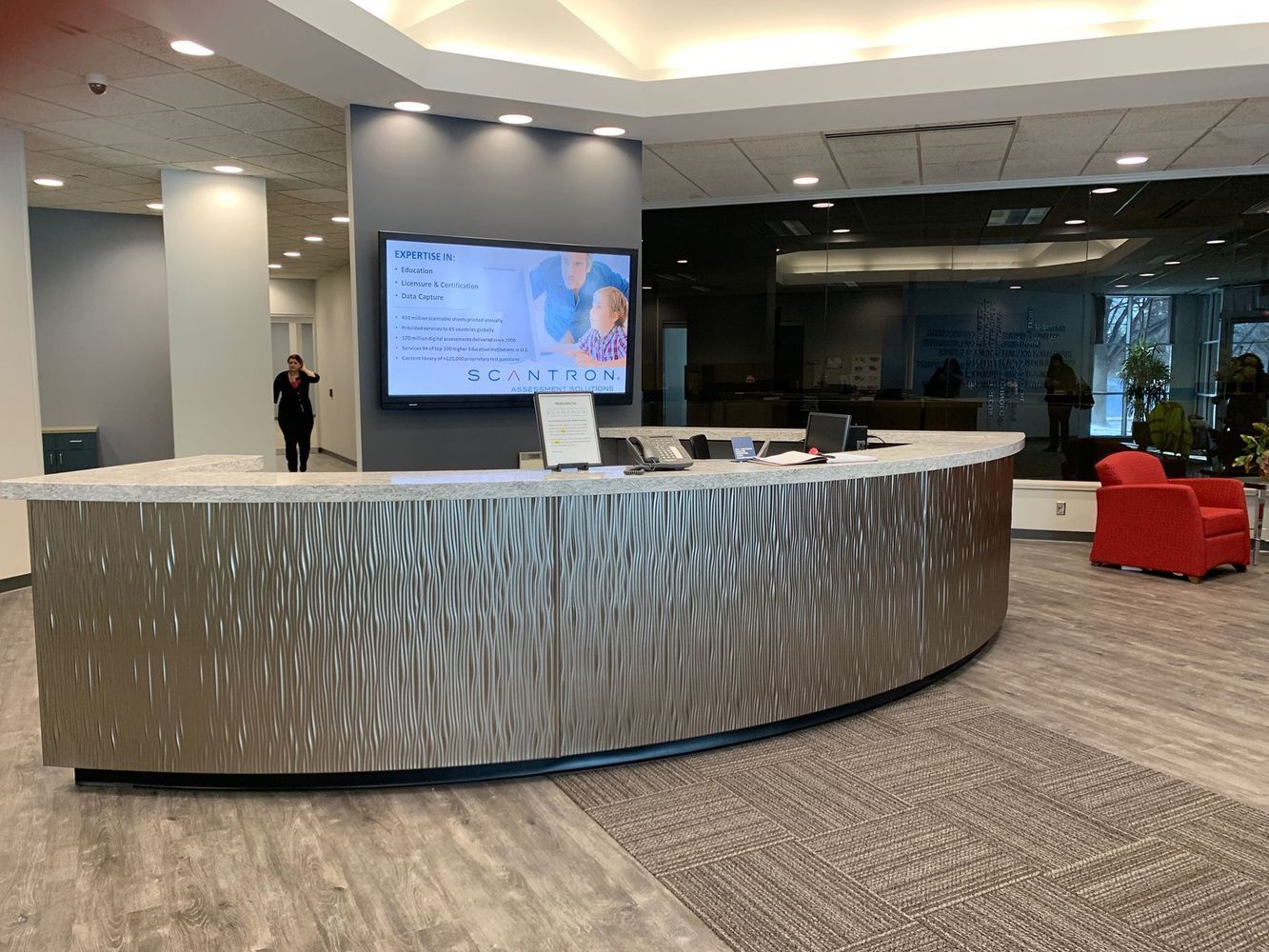 Reception desk with a curved, textured metal facade; a screen displays information. A person stands behind the desk.