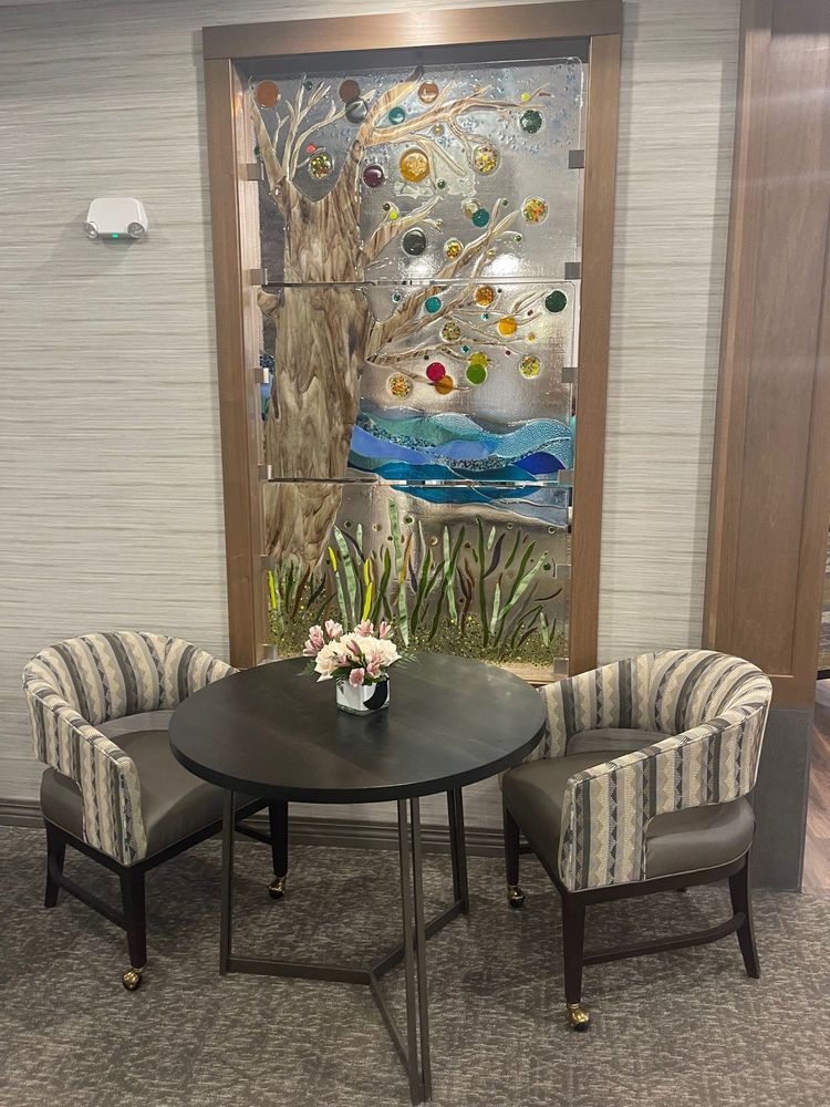 Two armchairs facing a round table. A stained glass panel of a tree and landscape is in the background.