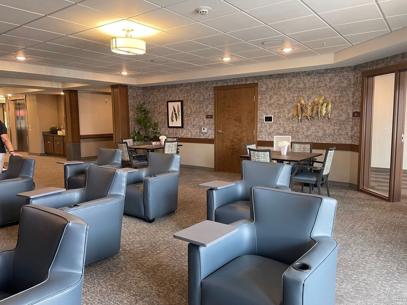 A room with blue armchairs, small tables, and round dining tables. Brown patterned wallpaper. Carpeted floor.