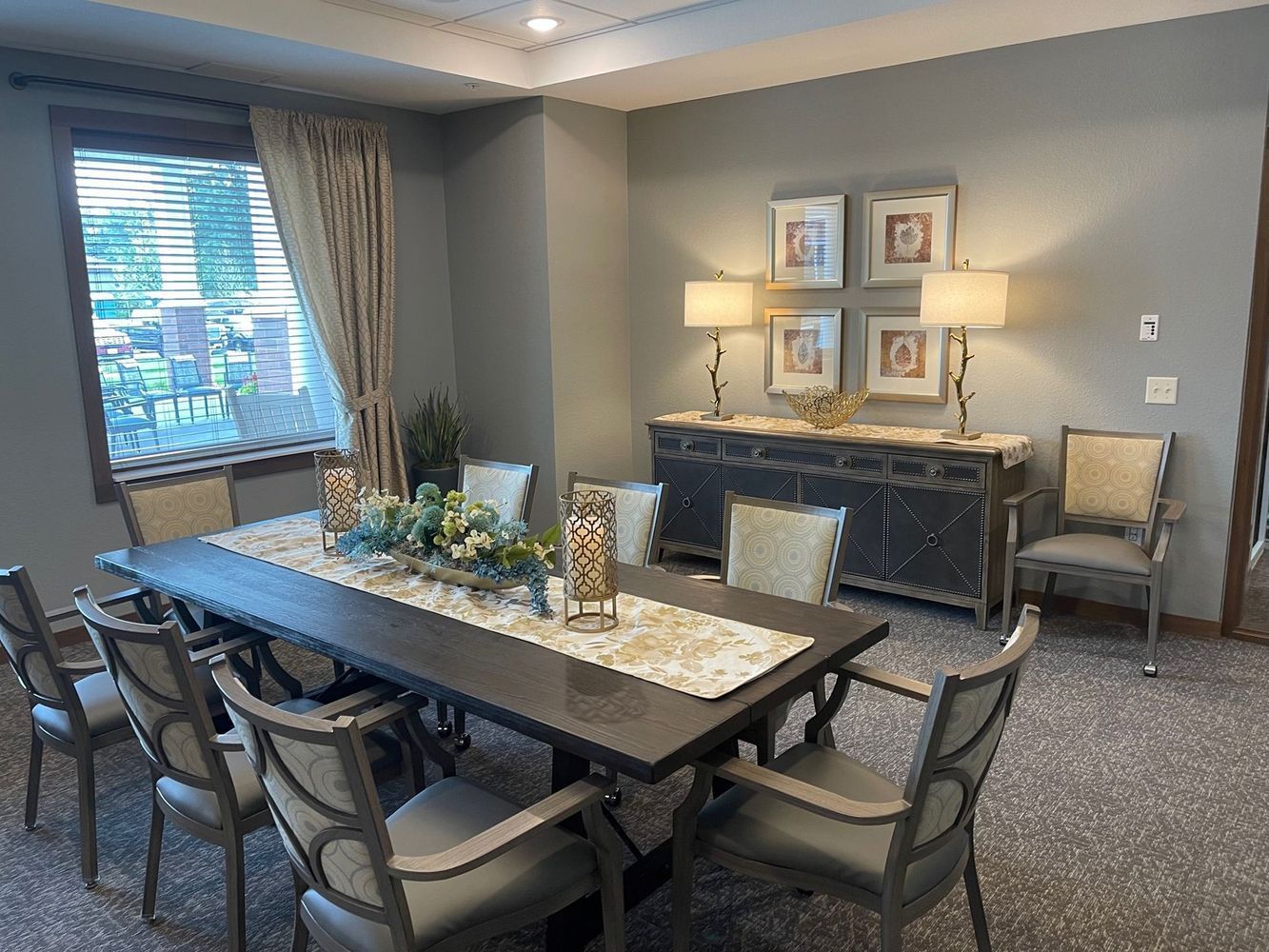 Formal dining room with long table, buffet, lamps, framed art, chairs, and window with curtains.