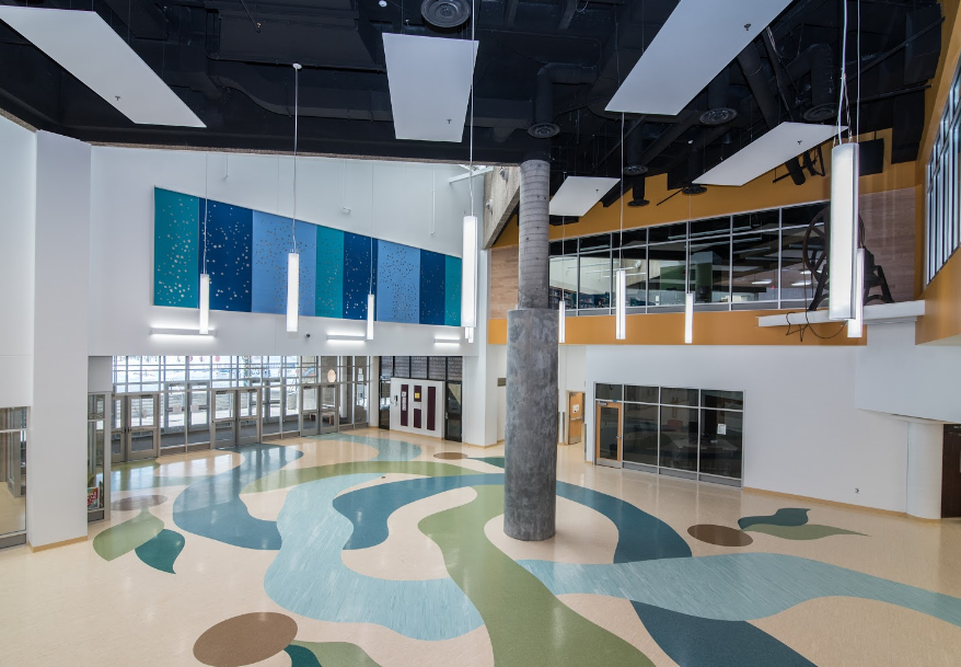 Large, open interior with abstract floor design and tall column. Blue and green accents, black ceiling with light fixtures.