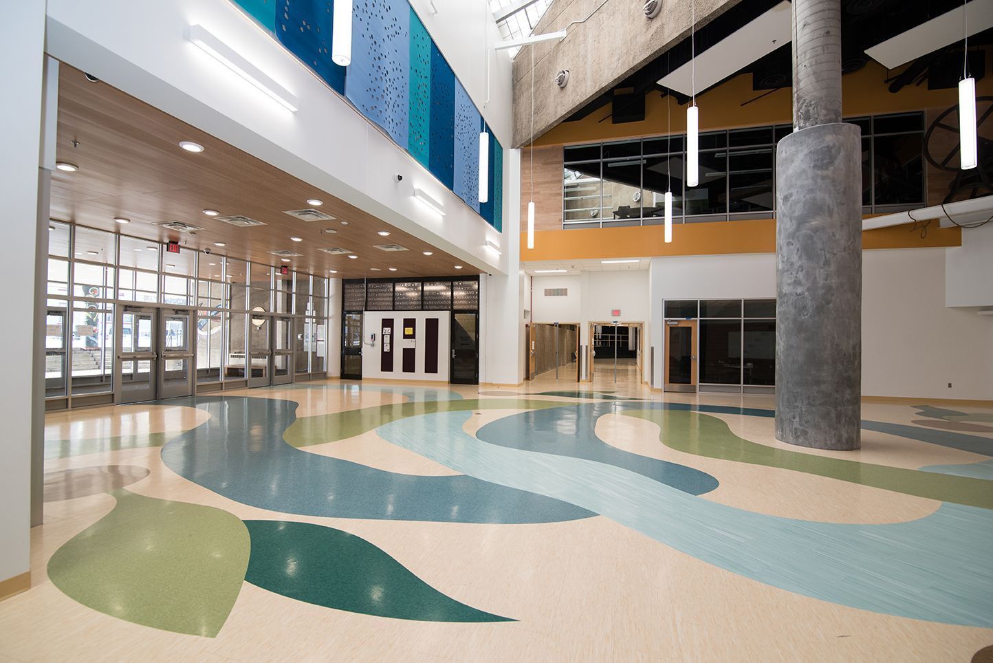 Spacious lobby with patterned floor, glass walls, a tall column, and a colorful mosaic wall.