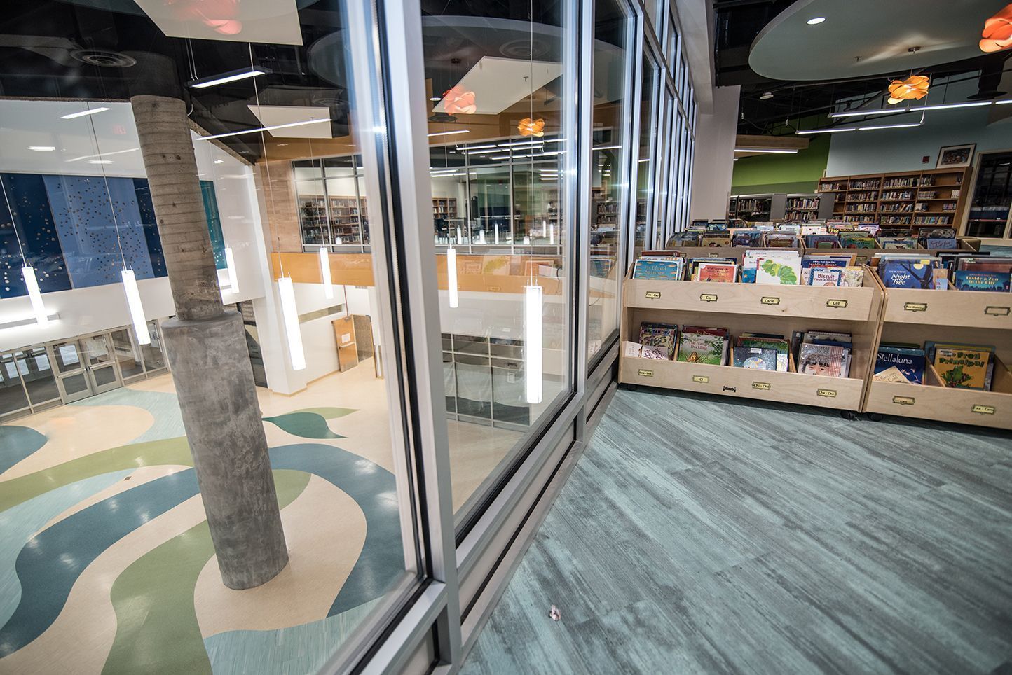 Interior of a modern library, showcasing bookshelves, large windows, and colorful flooring, a support beam looks like a palm tree.