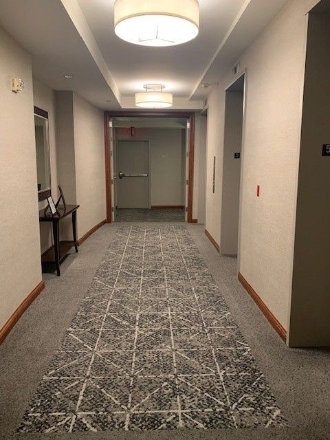 Hallway with gray patterned carpet, beige walls, and overhead lighting leading to an exit.