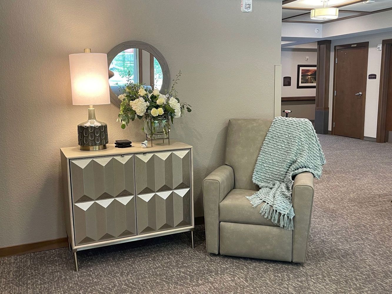 A cozy seating area in a hallway with a cabinet, lamp, mirror, chair, and blanket.