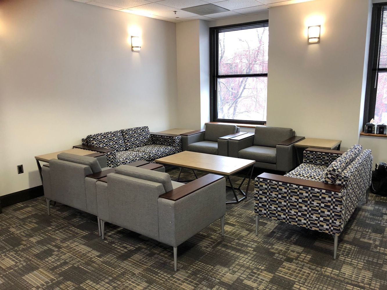 Lounge area with gray sofas, patterned chairs, small tables, a window, and wall sconces.