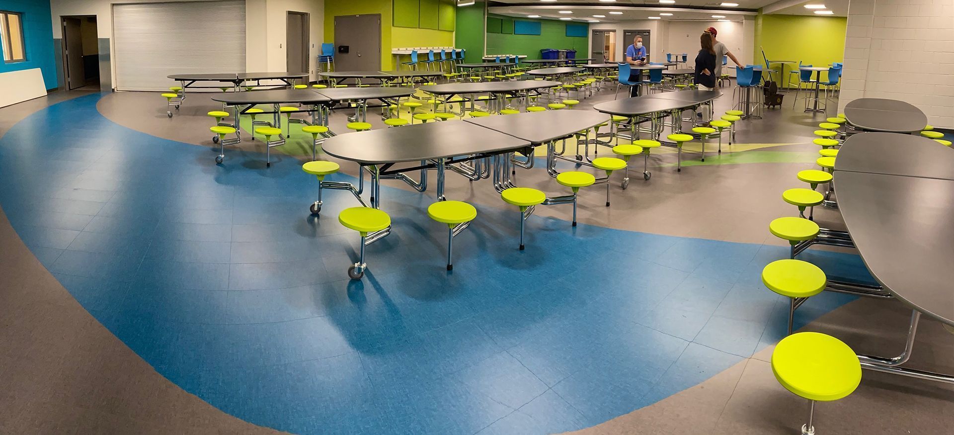 School cafeteria with tables and yellow stools, blue and gray flooring, and people walking around.