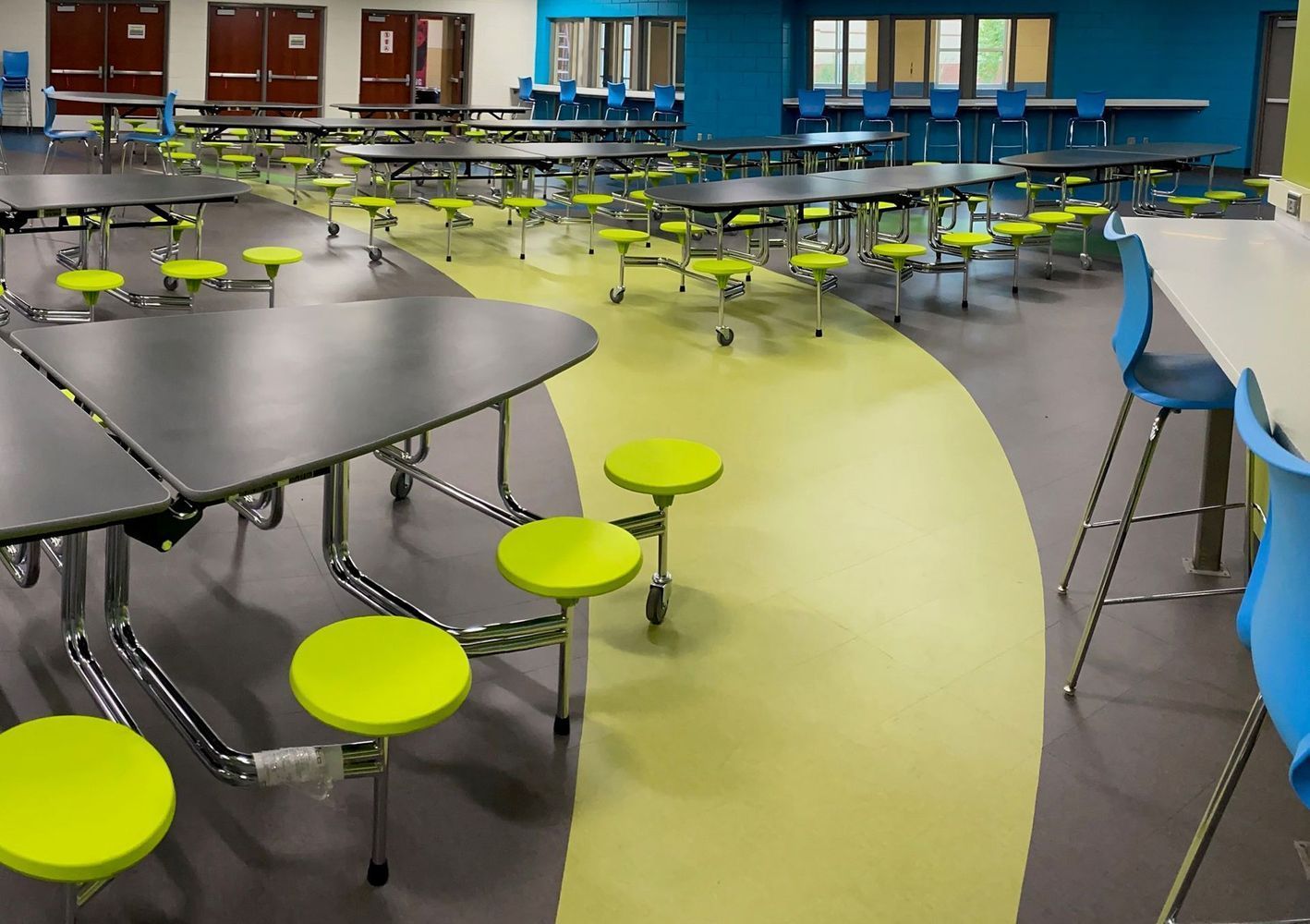 School cafeteria with green and gray floor, tables, and lime green stools. Blue wall and chairs are also present.