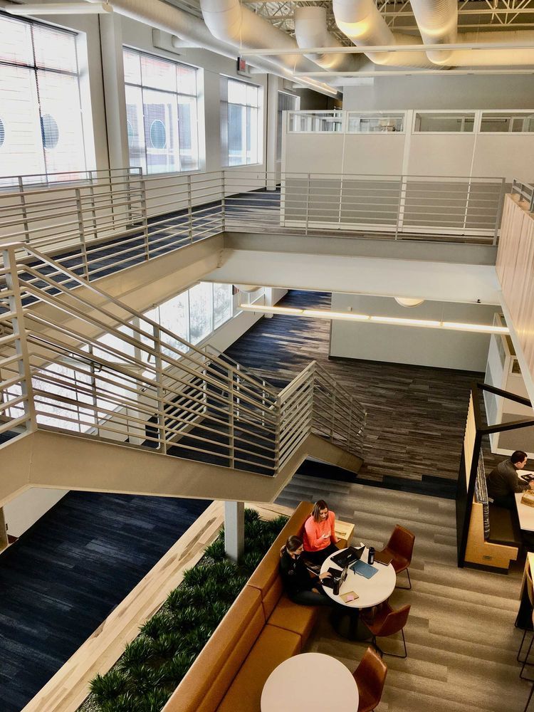 Multilevel office interior with stairs, walkway, and a person seated at a table. Blue, gray, and tan.