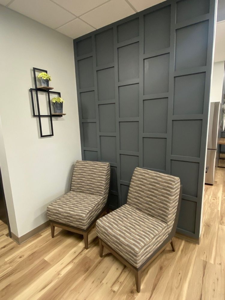 Two chairs in a waiting area with gray panel wall and wooden floors. A shelf with flowers on the left.