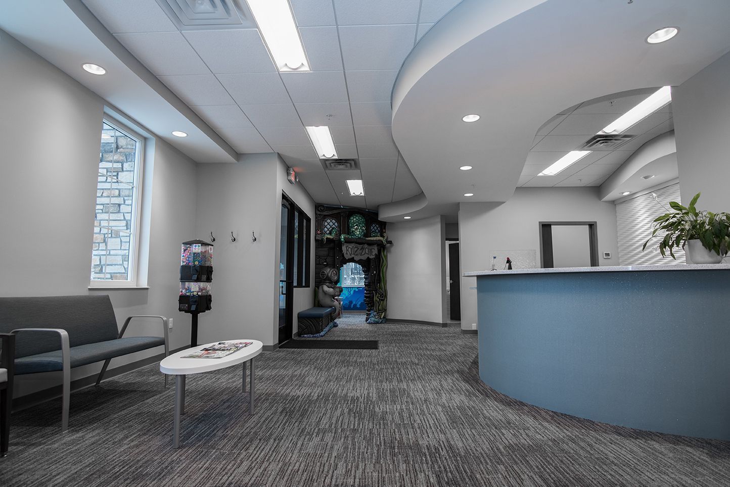 Waiting room with a gray sofa, reception desk, and water dispenser. Carpeted floor and neutral walls.