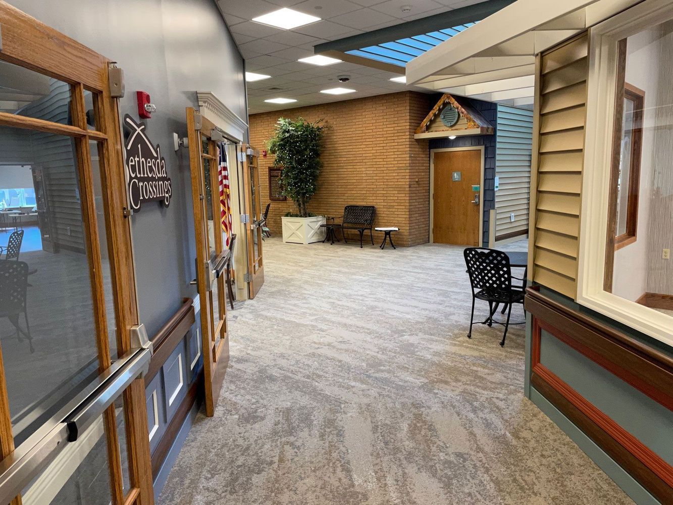 Hallway with doors, brick accent wall, and a large plant. Carpeted floor and overhead lighting.