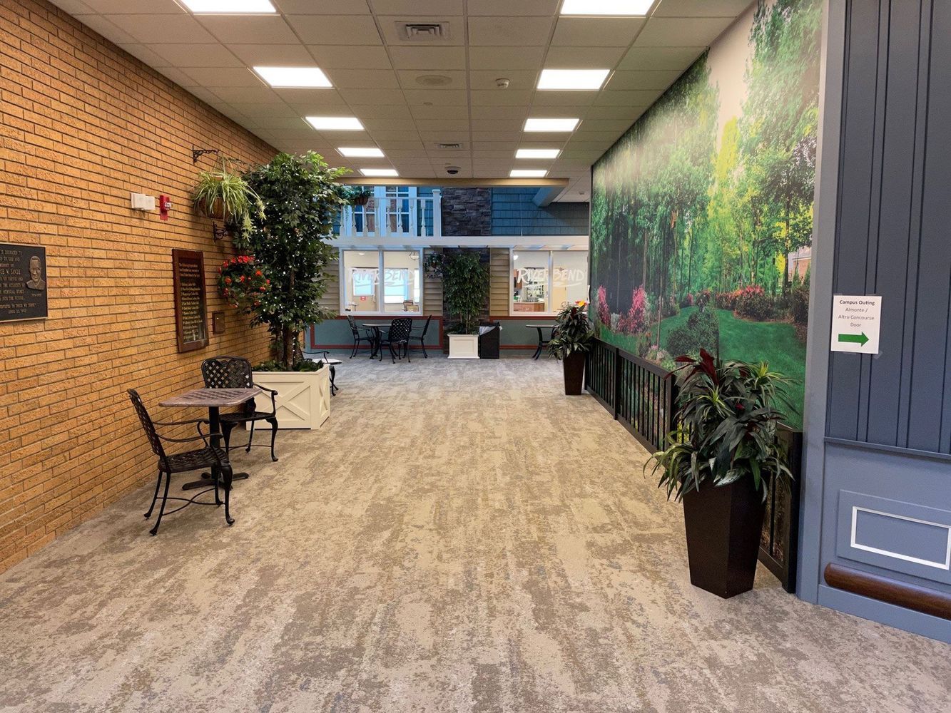 Hallway with brick wall, mural, tables, and plants; leading to a wider space with more tables and windows.