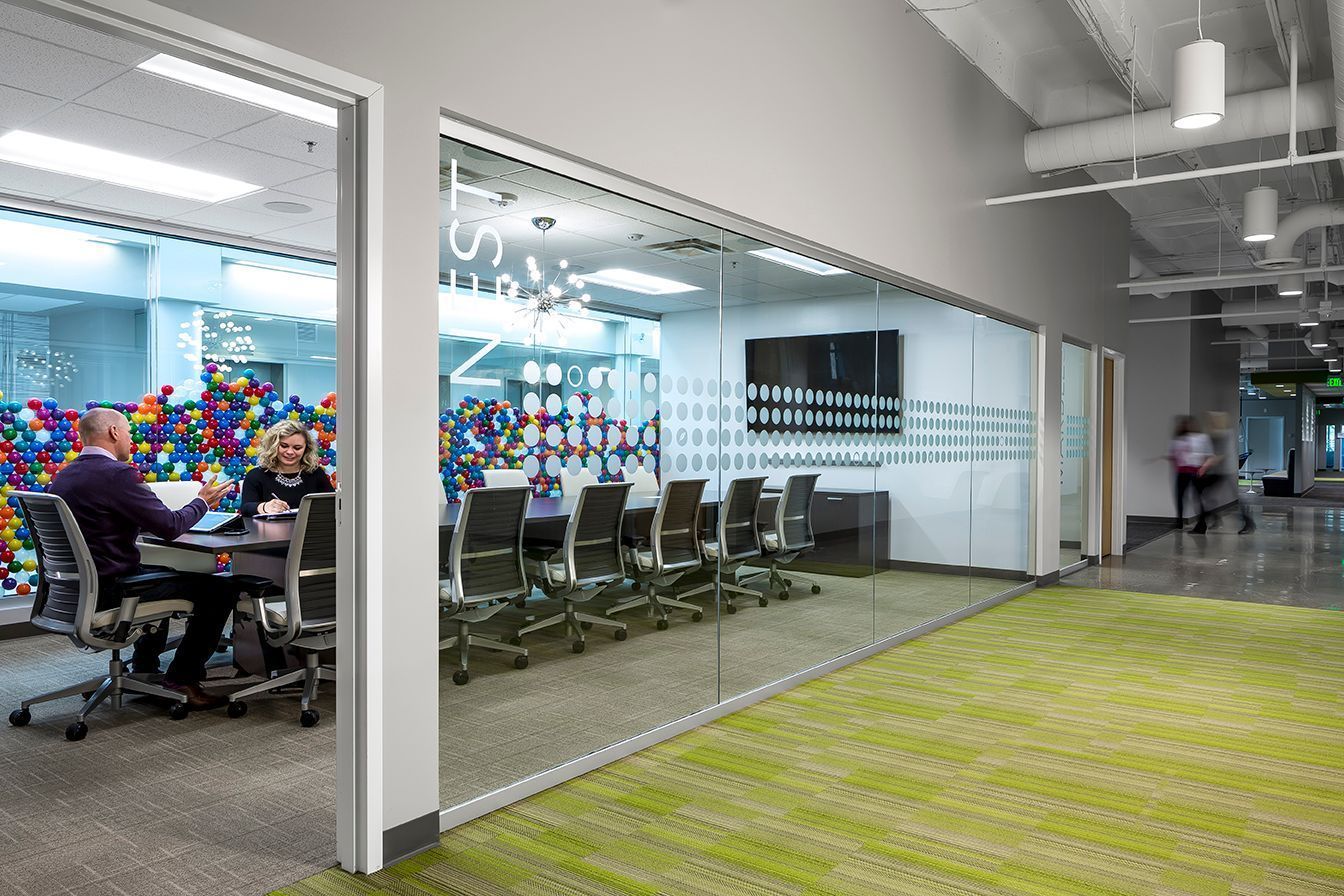 Office conference room with glass walls; people at table; green carpet; person walking in hallway.