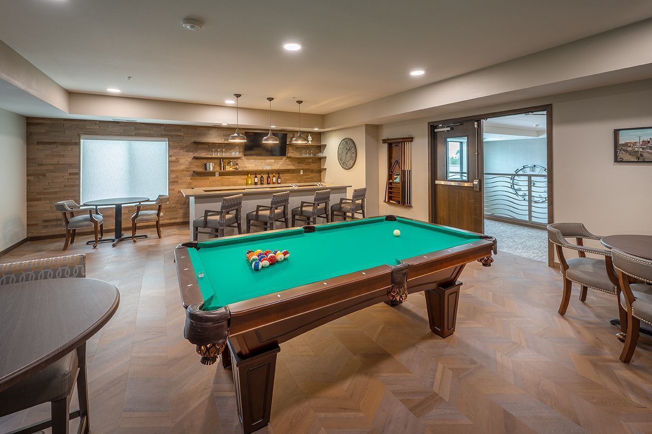 Pool table in a room with a bar, chairs, and tables. Wooden floors and walls; neutral colors.