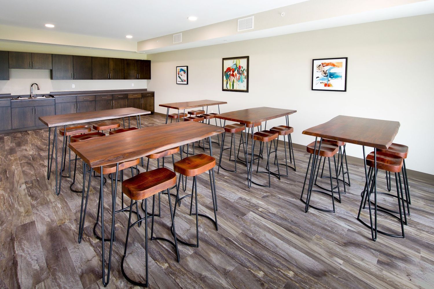Room with wooden tables, stools, and artwork. A kitchenette is in the background.