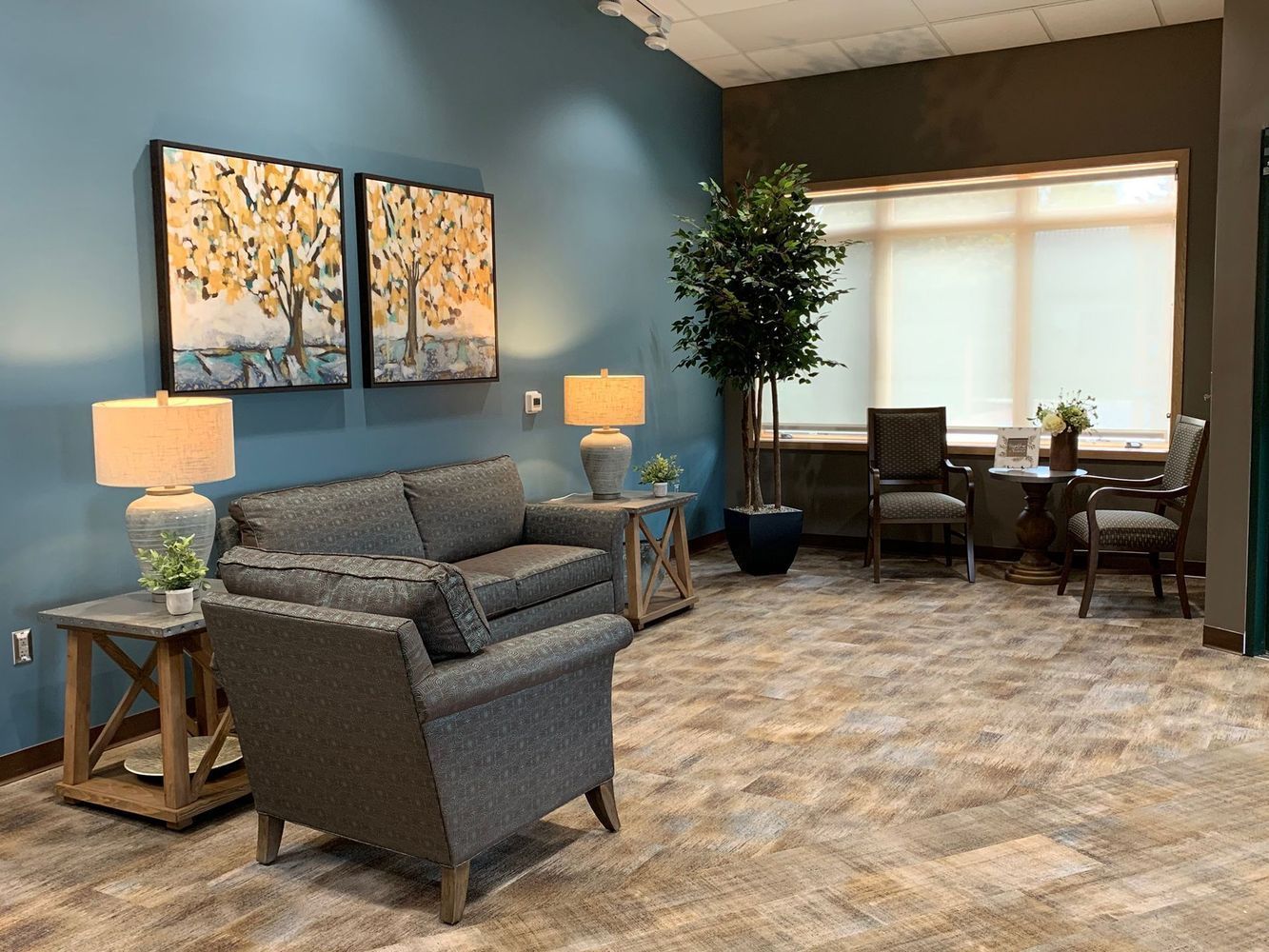 Cozy waiting room with blue wall, artwork, sofa, chairs, lamps, and a large tree next to a window.