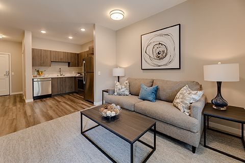 Living room and kitchen, neutral tones. Sofa, coffee table, artwork. Light wood floors and cabinets.