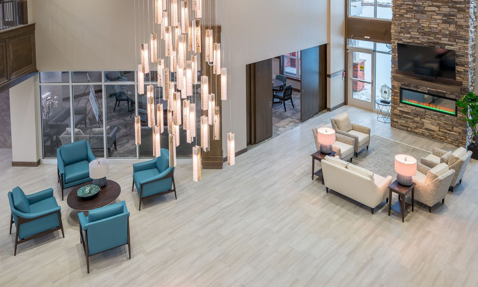 Lobby with seating areas: teal armchairs around a coffee table, cream sofas, fireplace, tall windows, and a large chandelier.