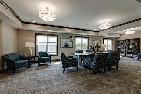 Cozy lounge with armchairs, table, lamps, and bookshelves. Neutral colors, large windows, and decorative lighting.