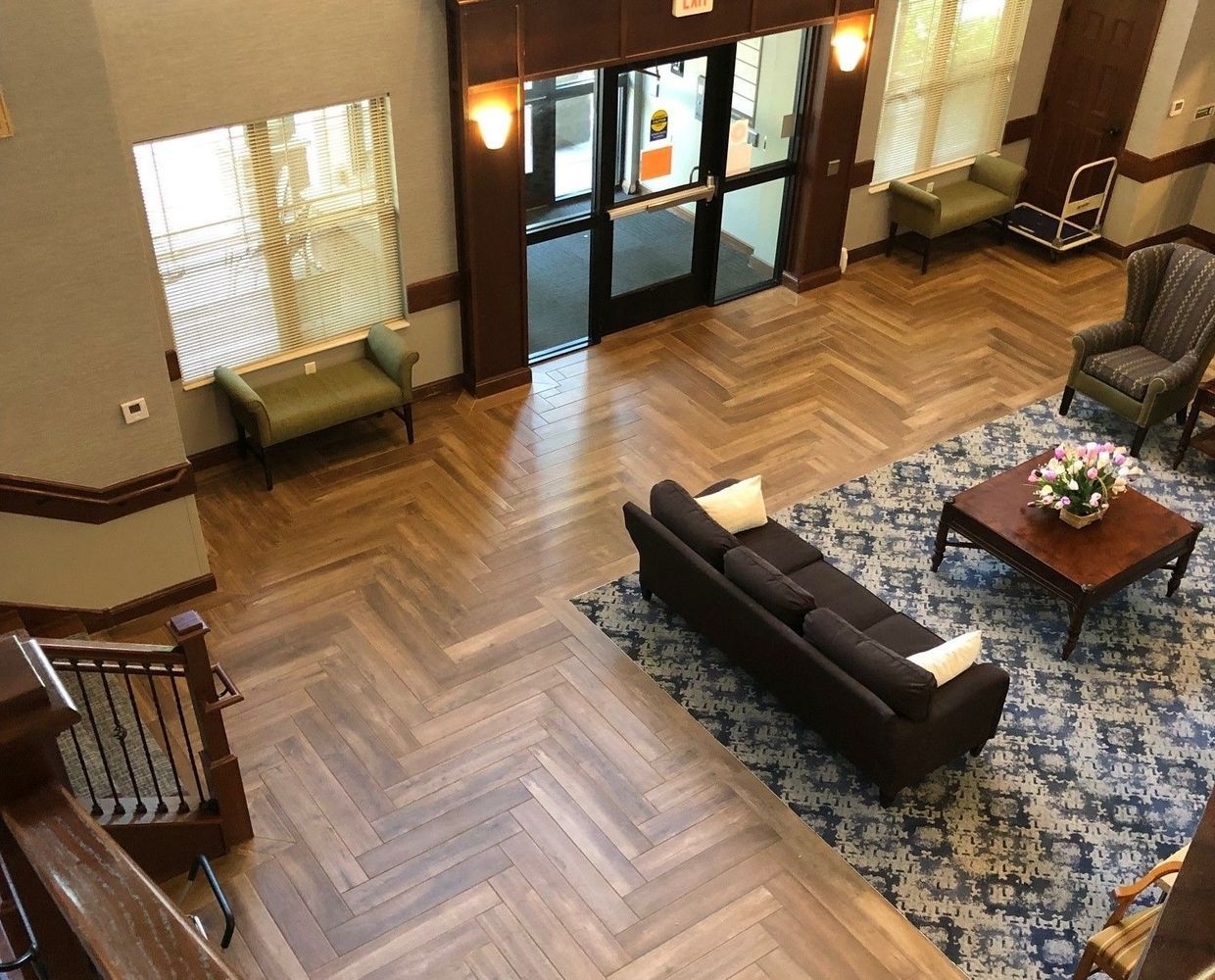 Lobby with wooden floors, seating areas, and a double-door entrance. Light streams in from windows.