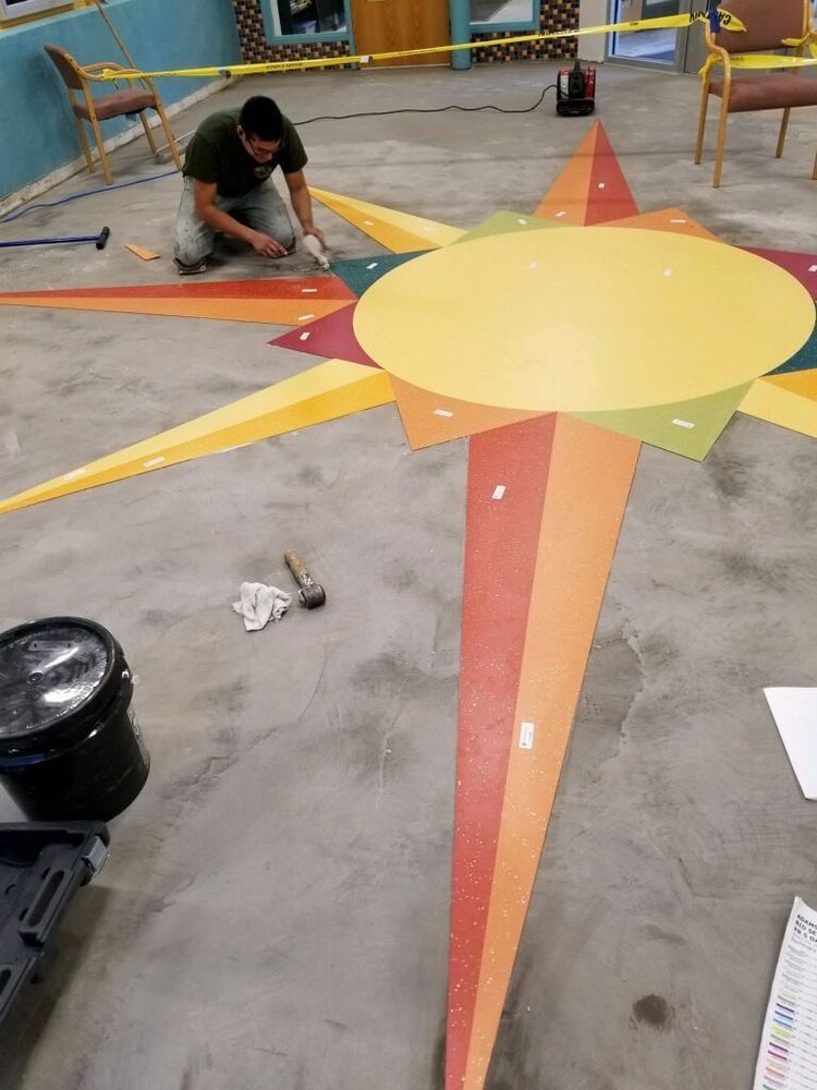 Person installs colorful starburst design on a concrete floor using a mallet, brush, and other tools.
