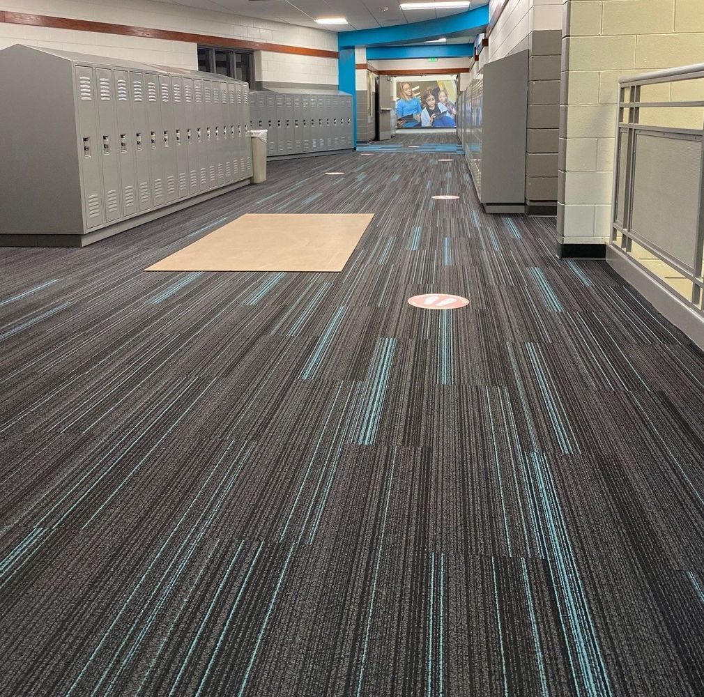School hallway with gray lockers, patterned carpet, and social distancing markers.