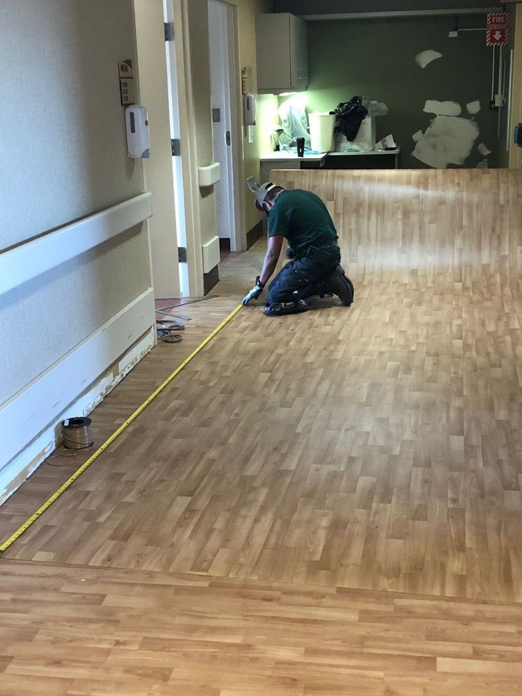Person kneeling, measuring floor in hallway with wood-look flooring, door frame visible.