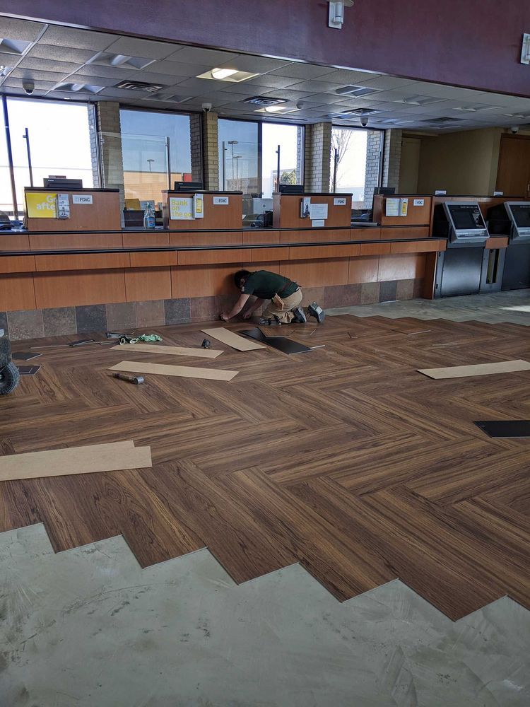 Bank interior, worker installing herringbone wood-look flooring. Brown and tan tones dominate.