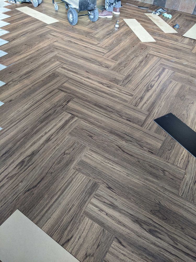 Herringbone wood-look flooring being installed in a room. Brown planks arranged in a diagonal pattern.