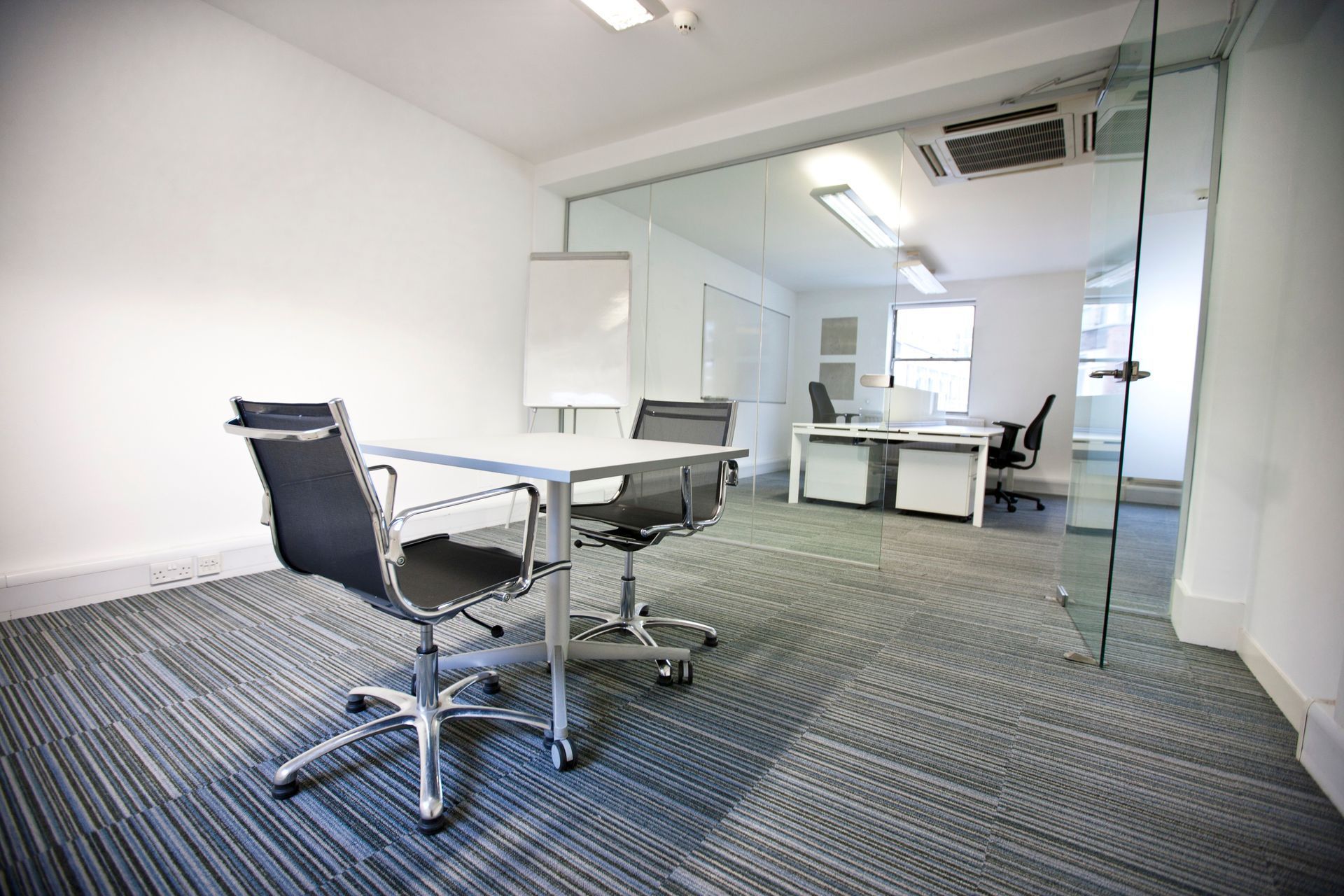 Empty office with a table, chairs, whiteboard, and a view into a workspace with desks.
