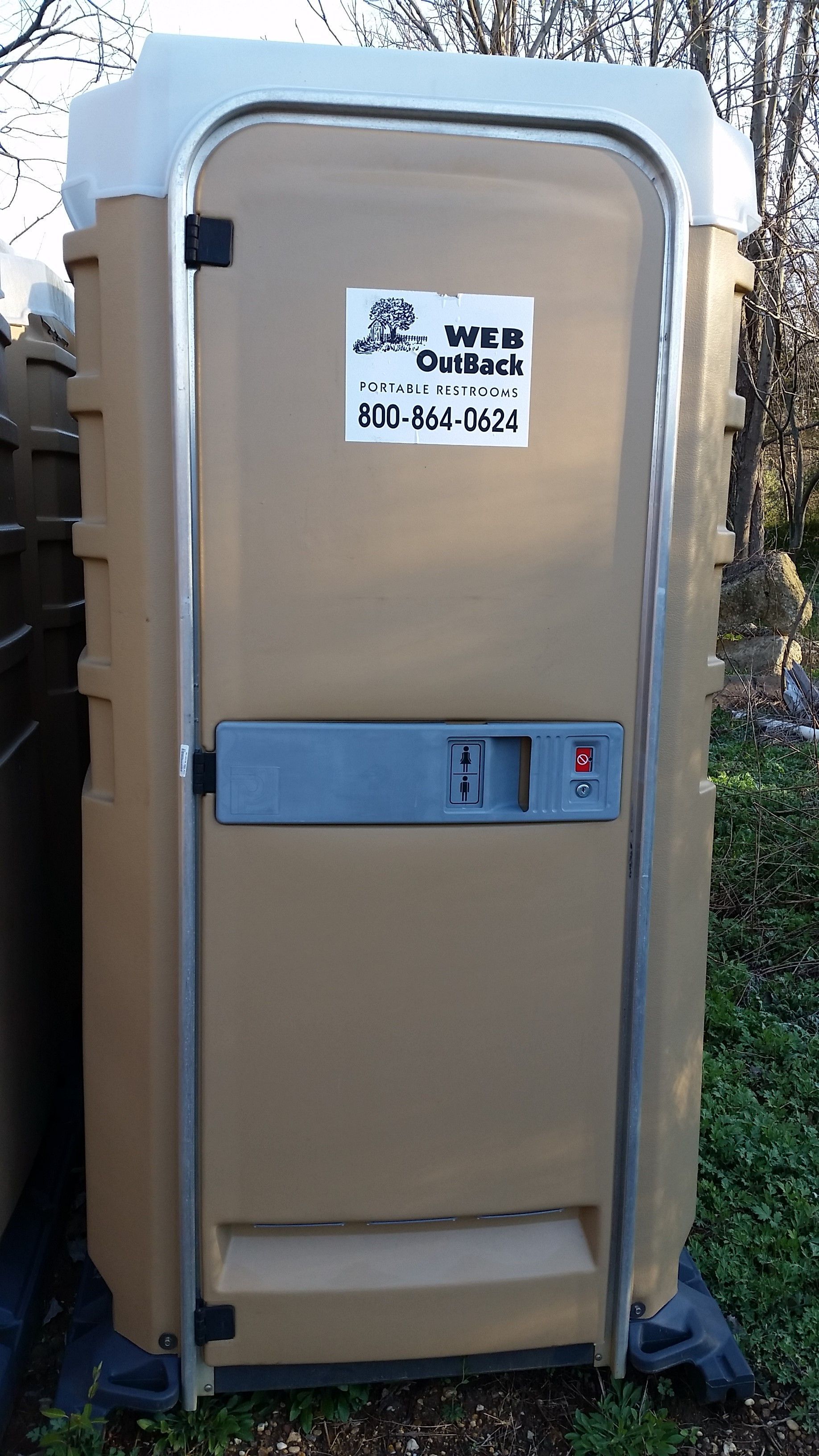 Tan portable toilet with a blue door handle and a sign on the front, outdoors.