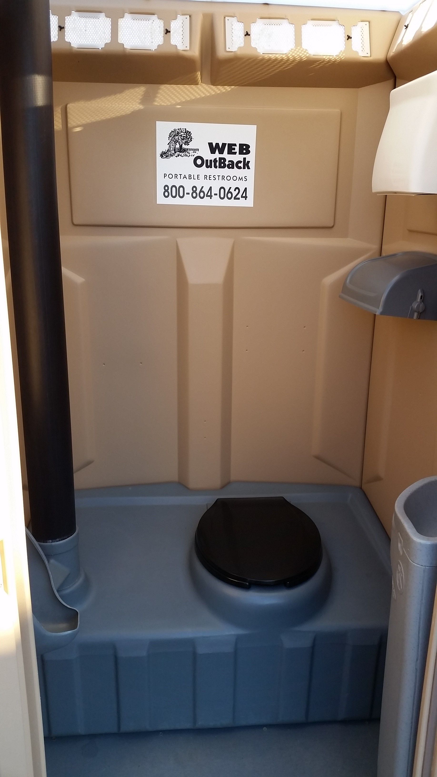 Portable outdoor toilet with tan walls and blue floor, featuring a black toilet seat and small sign above.